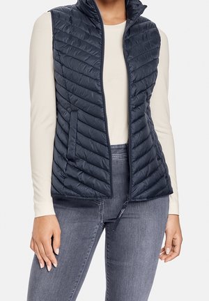 Kobieta ubrana w granatową pikowaną kamizelkę bez rękawów na kremowej bluzce z długim rękawem i szarych dżinsach, stojąca z rękami swobodnie opuszczonymi wzdłuż ciała.