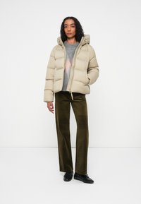 Chaqueta acolchada beige con capucha, con cierre de cremallera y detalles elásticos. Combinada con pantalones de pana verdes y zapatos negros.