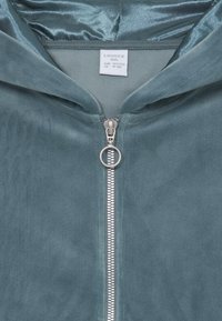Hettejakke i myk teal-farge med en glatt tekstur, med en metallglidelås og en løkke for å dra. Lappen viser størrelsesdetaljer.