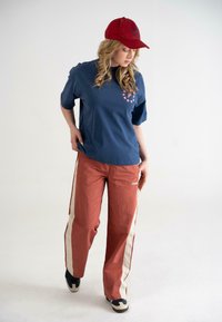 Marineblå bomulds T-shirt med lyserødt stjerneprint og logo, parret med rustfarvede bukser med hvide sidestreger, båret med sorte sneakers.
