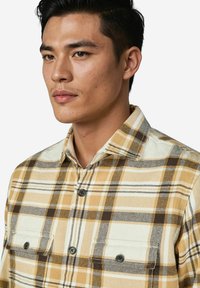 Junger Mann mit kurzen, dunklen Haaren, der ein beige-braun-weiß kariertes Hemd mit zwei Brusttaschen trägt.