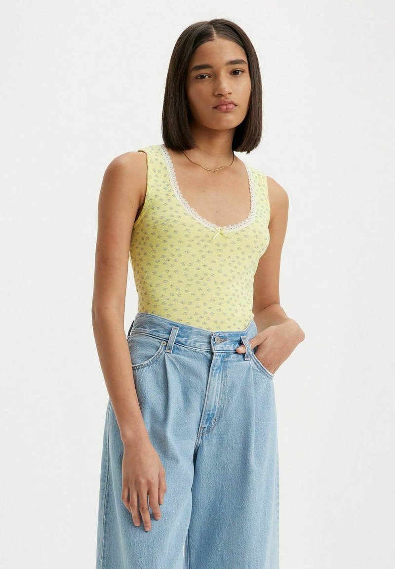 Bodysuit amarillo sin mangas con un patrón floral, detalles de encaje en el escote, combinado con jeans de tiro alto en un tono azul claro.