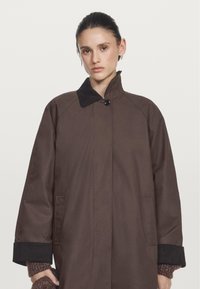 Brown oversized coat with a high collar, button closure, and large pockets. Features contrasting black cuffs and a smooth, matte finish.