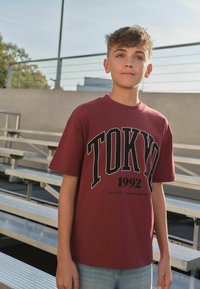 Jugendlicher mit kurzen Haaren, der ein dunkelrotes "TOKYO 1992" T-Shirt und Jeans trägt, steht draußen neben leeren Metall-Bankreihen.