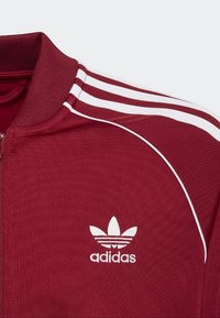 Veste zippée bordeaux avec accents de trois bandes blanches sur les épaules et un logo brodé blanc sur le côté gauche de la poitrine. Tissu texturé.