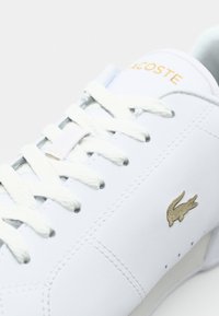 Baskets blanches Lacoste avec lacets blancs, inscription Lacoste dorée sur la languette, et petit logo crocodile doré sur le panneau latéral.