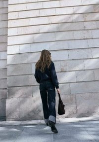 Czarny sweter i dżinsy z wysokim stanem, szerokie nogawki z jasnym mankietem; trzyma brązową torbę. Stoi w pobliżu kamiennej ściany o różnorodnych fakturach.