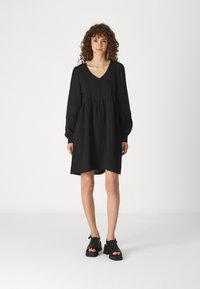 Vestido preto de mangas longas com decote em V, detalhes em botão e cintura franzida. Combinado com sapatos de plataforma pretos, textura suave e design casual.