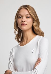 Biała koszulka z długim rękawem wykonana z gładkiej tkaniny, z małym czarnym logo na lewej piersi i okrągłym dekoltem.