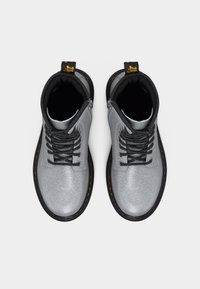 Ζευγάρι μεταλλικά ασημί μποτάκια με κορδόνια, μαύρες σόλες, μαύρα επενδεδυμένα κολάρα και κίτρινες θηλιές έλξης, όπως φαίνονται από πάνω σε λευκό φόντο.