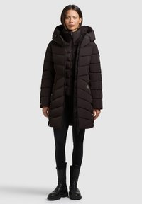 Schwarze Puffermantel mit Kapuze, mit Reißverschluss, gestepptem Design und elastischen Bündchen. Trägt man über schwarzen Leggings und klobigen schwarzen Stiefeln.