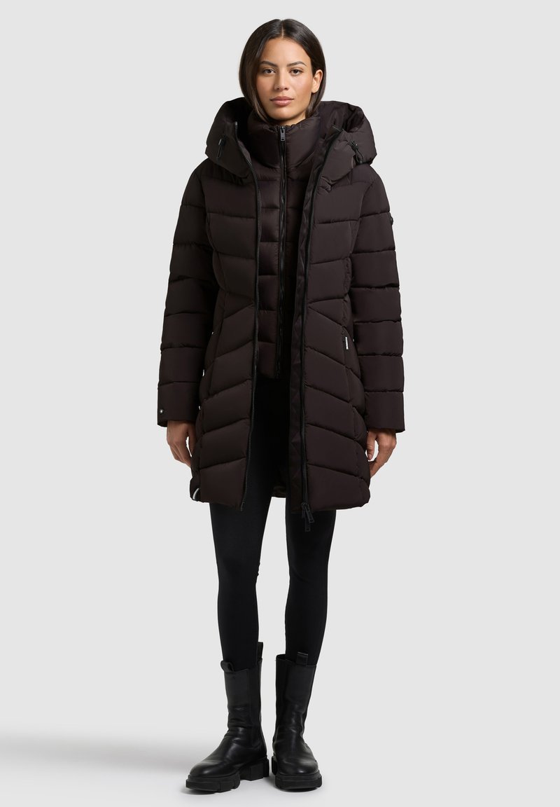 Schwarze Puffermantel mit Kapuze, mit Reißverschluss, gestepptem Design und elastischen Bündchen. Trägt man über schwarzen Leggings und klobigen schwarzen Stiefeln.