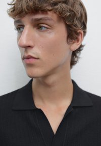 Polo nero con tessuto testurizzato, dotato di colletto e vestibilità slim, indossato da una persona con pelle chiara e capelli arruffati.