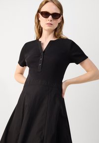Black dress featuring a ribbed top with a henley neck and short sleeves, transitioning to a smooth, flared skirt design with side pockets.