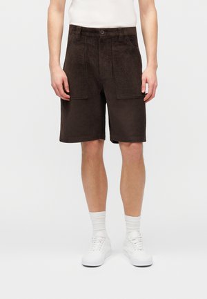 Άνδρας που φοράει σκούρα καφέ βερμούδα από κοτλέ μέχρι τα γόνατα, λευκές κάλτσες και λευκά αθλητικά παπούτσια μπροστά σε απλό φόντο.