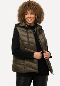 Olivově zelená pufová vesta s kapucí, s zipem vpředu a vertikálním prošíváním. Nahoře má černý svetr s tkaničkami a kapsami.