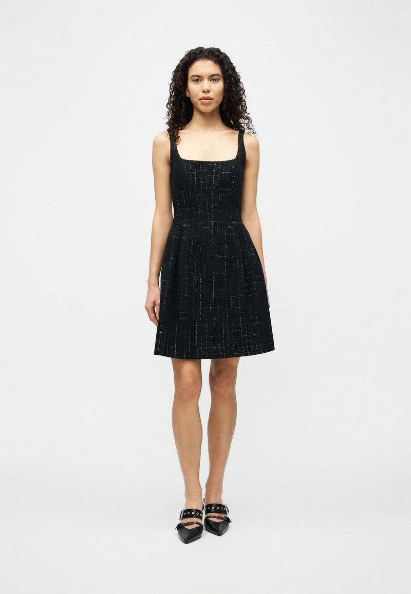 Γυναίκα που στέκεται με το πρόσωπο προς τα εμπρός, φορώντας αμάνικο μαύρο φόρεμα με διακριτικές ασημένιες γραμμές και μαύρα μυτερά παπούτσια με λουριά.