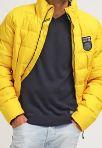 Doudoune jaune avec un motif matelassé et une fermeture éclair noire. Portée sur un pull bleu marine, avec un écusson logo circulaire. Jean bleu clair.