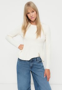 Cream ribbed sweater with long sleeves, round neckline, and a flared hem, paired with high-waisted blue wide-leg jeans.