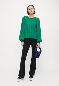 Green ribbed long-sleeve top with puffed sleeves, paired with black flared pants and a navy knitted handbag. White sneakers complete the look.