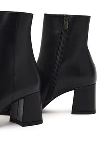 Bottines en cuir noir avec un talon carré, détail de fermeture éclair sur le côté, texture lisse et design minimaliste.