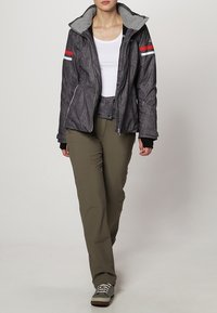 Giacca grigia strutturata con cappuccio grigio, strisce rosse e bianche sulle maniche, abbinata a pantaloni verde oliva e sneaker grigie.