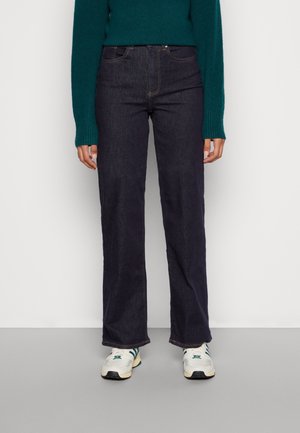 Personne portant un jean droit bleu foncé, un pull vert à manches longues et des baskets blanches avec des bandes vertes, debout sur un sol gris.
