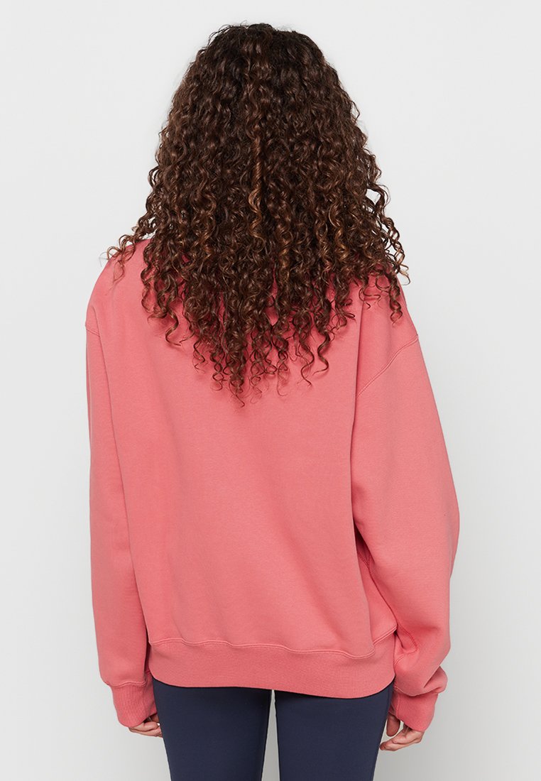 Osoba z długimi kręconymi brązowymi włosami, ubrana w luźny różowy sweatshirt i ciemne legginsy, widziana od tyłu na jednolitym tle.