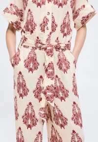 Femme portant une combinaison beige avec un motif floral paisley rouge, des boutons sur le devant, une ceinture en tissu nouée et des manches courtes, mains dans les poches.