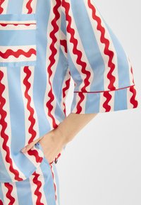 Kortærmet skjorte med bølgede røde og hvide striber på en lyseblå baggrund, med brystlomme og kontrastdetaljer ved kanterne.