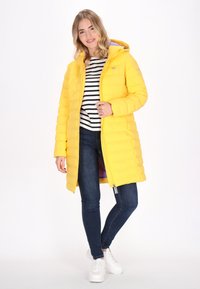 Gul puffjacka med huva, som har dragkedja, horisontell sömnad och lila foder. Bärs över en svart- och vitrandig skjorta.