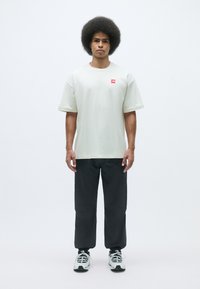 Light beige cotton t-shirt with short sleeves and a small red logo, paired with dark gray pants and white sneakers with black accents.