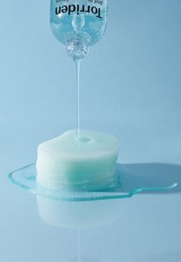 Klar blå gel hælder fra en omvendt flaske på en lys blå, rund, lagdelt gelbasis med en let glans og en lille pøl af væske.