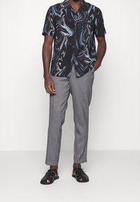 Uomo che indossa una camicia nera a maniche corte con spirali blu e bianche, pantaloni grigi e sandali di pelle nera intrecciati, in piedi con una mano in tasca.