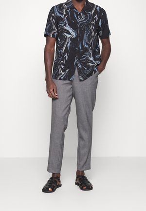 Hombre vestido con una camiseta negra de manga corta con remolinos azules y blancos, pantalones grises y sandalias de cuero negro trenzadas, de pie con una mano en el bolsillo.