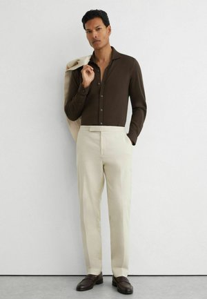 Mann trägt dunkelbraunes Hemd mit Knöpfen, off-white Hose, hält eine beige Jacke über der Schulter, steht an einer weißen Wand.
