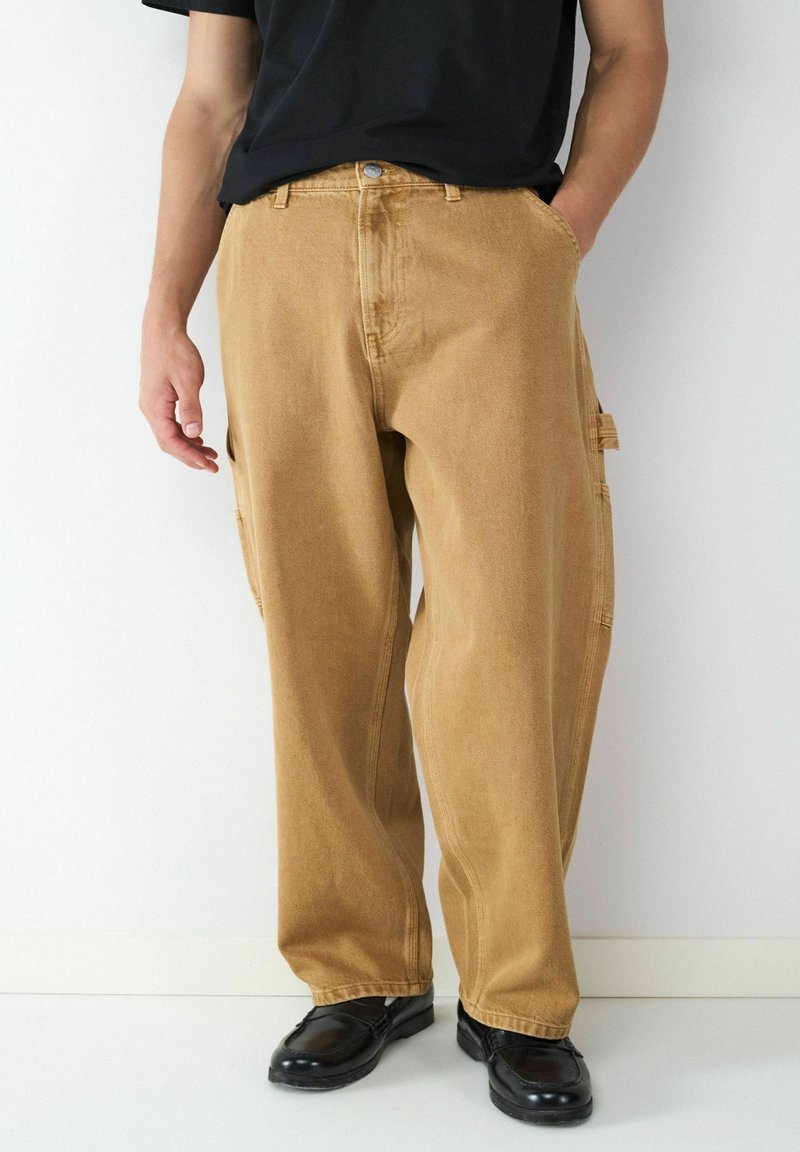 Beige Cargo-Hosen aus Denim, mit lässiger Passform, Seitentaschen und einem Reißverschluss mit Knopfverschluss. Getragen mit schwarzen Schuhen.