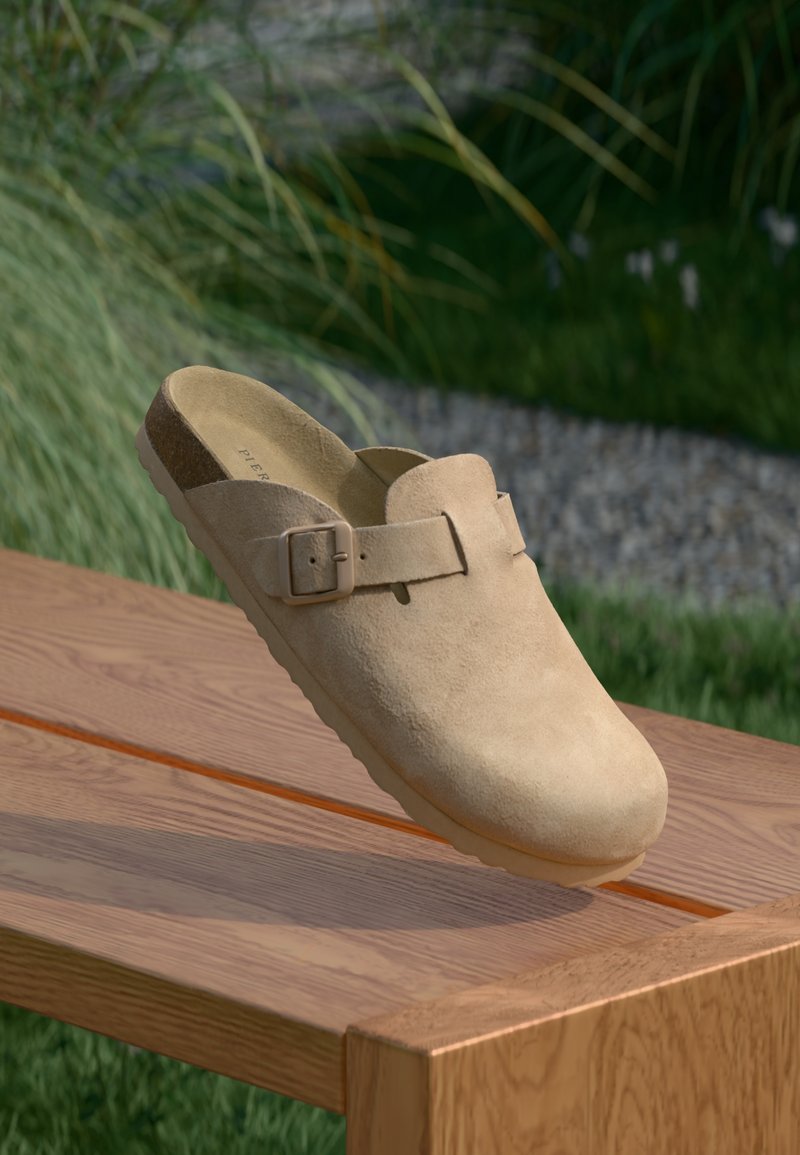 Bézs velúr klumpa lapos talppal, kerek orral és négyzet alakú csat kiegészítéssel. Fából készült felületen és zöld növényzet háttérben.