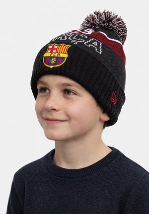 Young boy wearing a black and maroon knit hat with a pom-pom, featuring the FC Barcelona logo and text, smiling and looking slightly right.
