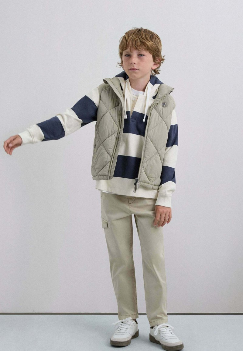Niño con chaleco acolchado beige, sudadera a rayas, pantalones beige y zapatillas blancas, de pie con un brazo extendido hacia un lado.