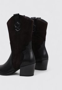 Botas de tobillo de cuero negro y gamuza oscura con tacón apilado, diseño curvado y acentos decorativos en la parte superior.