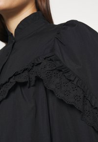Blouse noire avec col montant à volants et bordure en dentelle florale sur le yoke, détail d'épaule bouffante.
