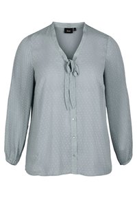 Blusa verde chiaro con scollo a V, dettaglio a nodo al colletto, maniche a pieghe verticali e piccoli motivi a pois bianchi.