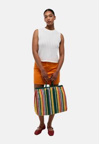 Sac fourre-tout rayé avec des lignes verticales rouges, vertes, jaunes et orange. Matière en tissu, forme rectangulaire, poignées vertes robustes.