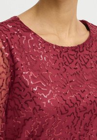 Zbliżenie na osobę w bordowej bluzce ozdobionej błyszczącymi, nieregularnymi falistymi wzorami z cekinów na przezroczystym materiale.