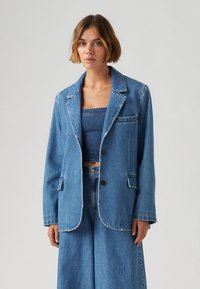 Denim blazer v svetlo modri barvi z ohlapnim krojem, zareznimi klapnicami in zapiranjem na en gumb. Ponuja sprednje žepe in detajle kontrastnega šivanja.