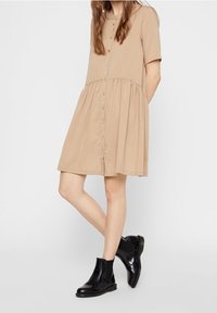 Abito beige con bottoni, maniche corte, gonna a strati e vestibilità rilassata. Indossato con stivali Chelsea neri. Tessuto morbido con una texture liscia.