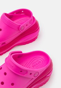 Des Crocs rose vif en matériau caoutchouteux, avec un design à enfiler, un talon surélevé, des trous de ventilation et une sangle pour un maintien sécurisé. Texture lisse.