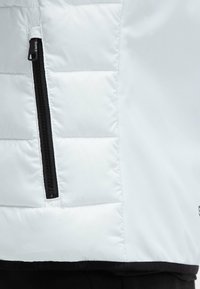 Sección de chaqueta blanca aislante con un patrón acolchado, que presenta un bolsillo con cremallera negra y un borde inferior negro suave. Detalle de tejido texturizado visible.