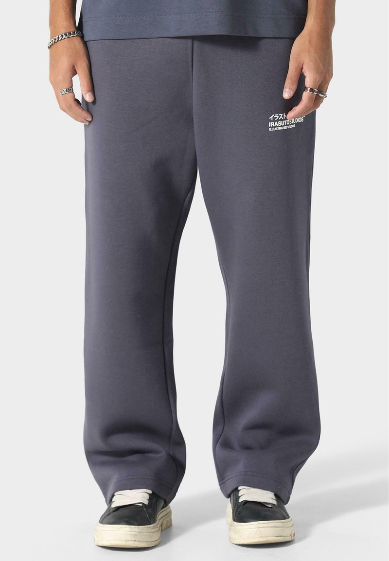 Grå joggingbukser med en afslappet pasform, der har en glat tekstur og detaljer i sidesømmen. Logo i hvid på venstre ben. Sorte sneakers med hvide snørebånd.